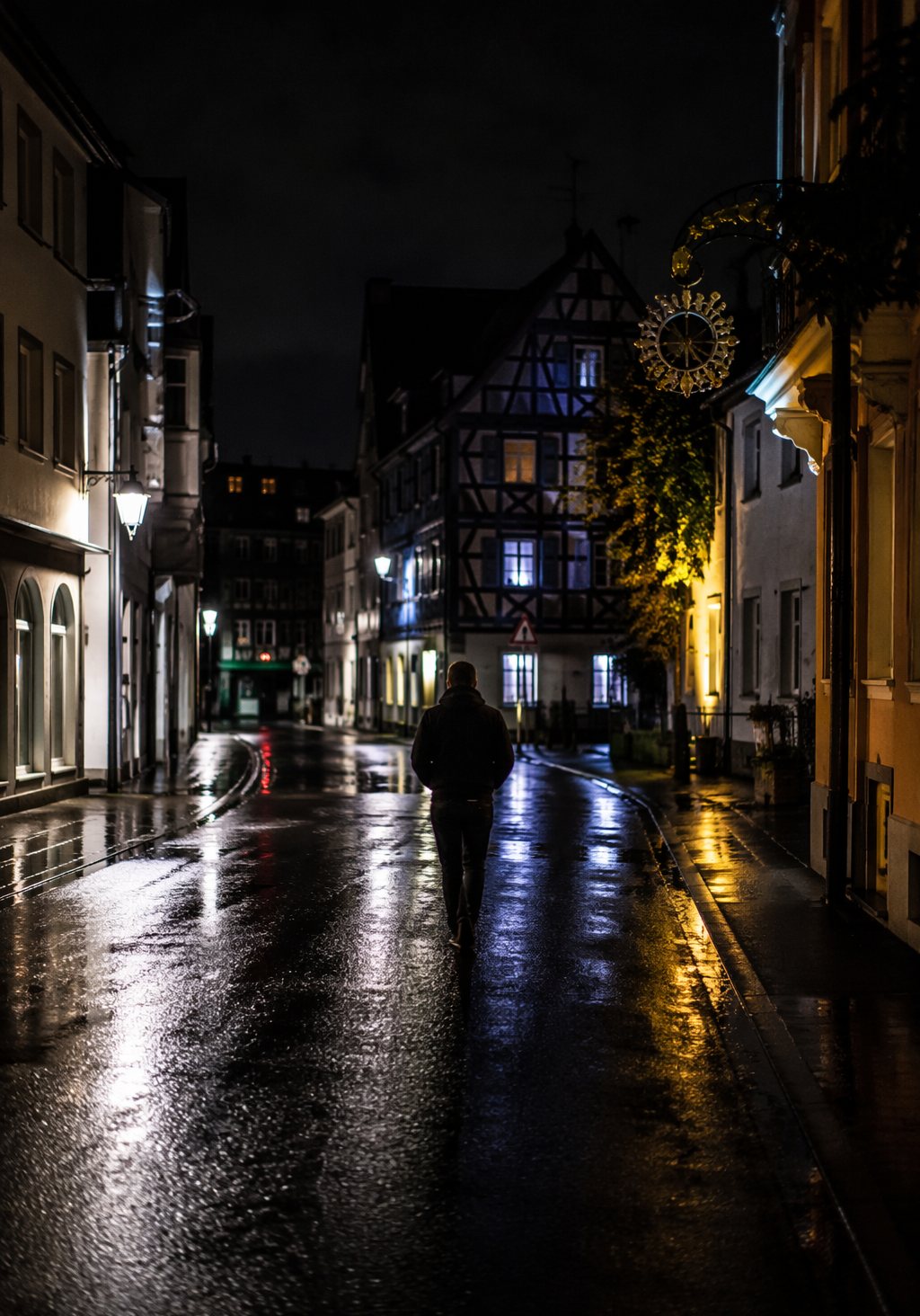Nächtliche Straße mit Fachwerkhäusern, nasser Asphalt spiegelt die Lichter, eine Person geht davon